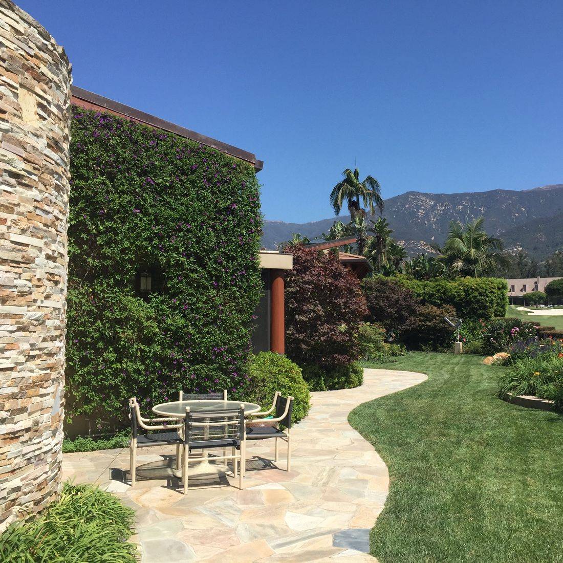A serene garden scene featuring a stone pathway leading to a small table and chairs surrounded by lush greenery and mountains in the background. The clear blue sky adds to the peaceful atmosphere.
