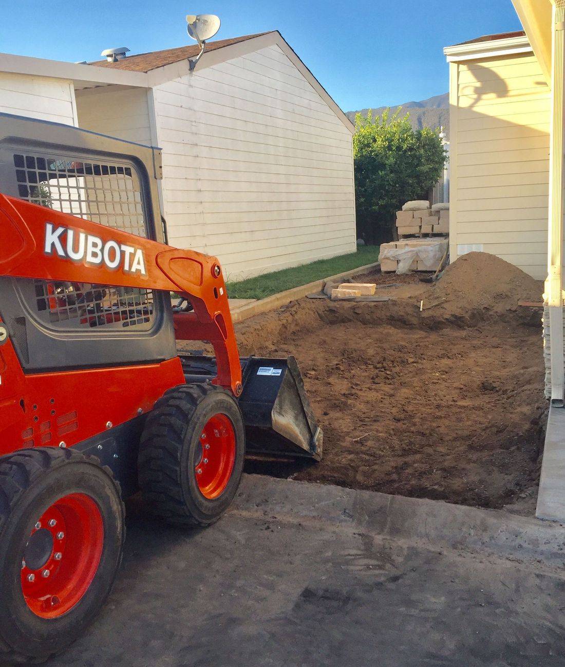 A compact construction vehicle is parked next to a freshly excavated area of dirt, with piles of soil and building materials visible in the background. The scene is set in a residential area with houses lining the sides.
