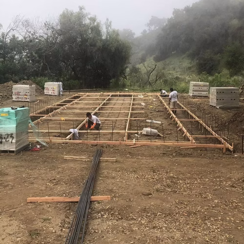 A construction site with workers preparing the foundation framework, surrounded by materials and a misty landscape.