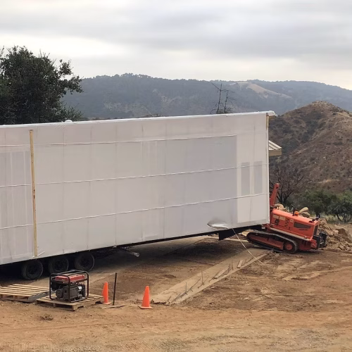 A large white structure is being transported on a construction site, with a mountainous landscape in the background. An orange vehicle is assisting in the movement of the structure.