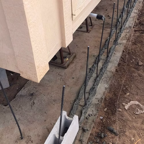 A construction site showing a foundation with exposed rebar and a concrete beam being prepared for a structure. The ground is partially excavated, indicating ongoing building work.