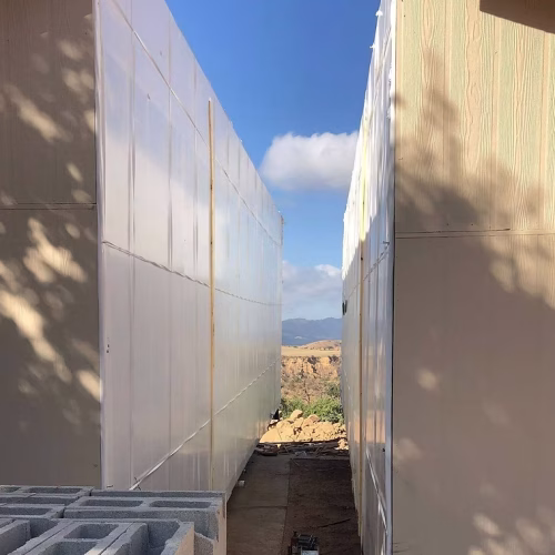 A narrow passage between two tall structures, with a clear blue sky and distant mountains visible in the background. The scene suggests a construction site or architectural development.
