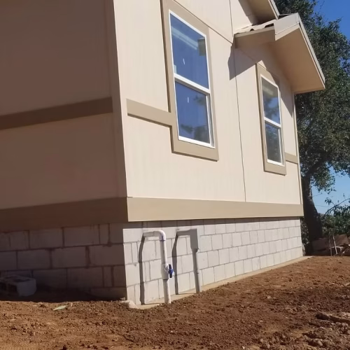 A newly constructed house with a light-colored exterior and multiple windows, situated on a dirt lot with exposed foundation. The image captures the base of the house, highlighting the construction details.