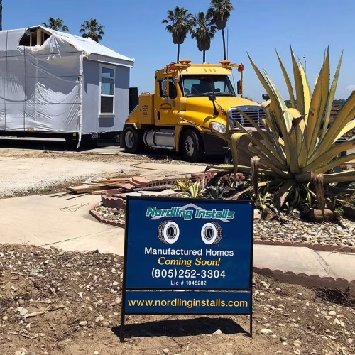 A yellow truck is parked next to a partially constructed manufactured home, with a sign advertising upcoming homes for sale. The setting includes desert landscaping with agave plants.