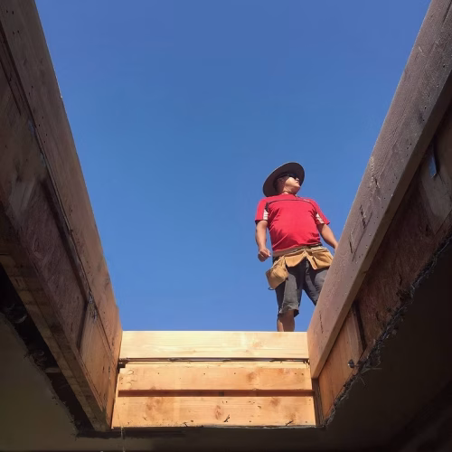A construction worker stands on a wooden structure, looking up at the clear blue sky. The scene captures the essence of manual labor and construction work.