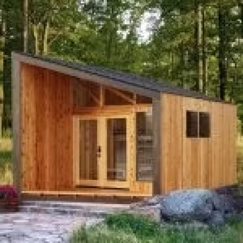 A small wooden cabin with a sloped roof is surrounded by trees, featuring large glass doors and a minimalist design. The cabin is set on a stone pathway, blending harmoniously with the natural environment.