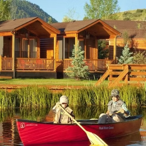 Two people are paddling a red canoe on a calm lake, with wooden cabins and greenery in the background. The scene captures a peaceful outdoor setting.