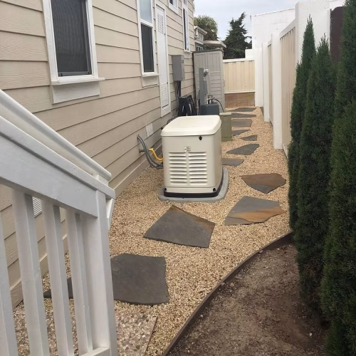 A narrow pathway lined with stepping stones leads to a generator beside a house, surrounded by gravel and shrubs. The setting is well-maintained and organized.