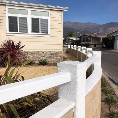 A modern house with a light-colored exterior is seen from the corner, featuring a white fence and landscaped garden. The backdrop includes mountains under a clear blue sky.