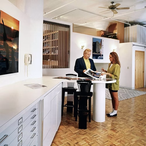 A man and a woman are engaged in a discussion at a modern, minimalist workspace, surrounded by architectural plans and designs. The space features wooden flooring and contemporary decor.