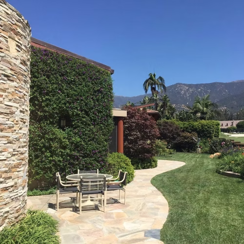 A stone wall covered in greenery leads to a patio area with a table and chairs, set against a backdrop of mountains and a clear blue sky. The landscape features well-maintained grass and various plants.