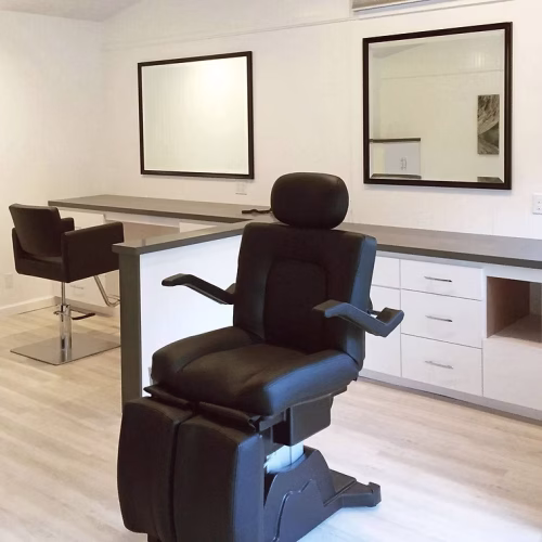 A black salon chair is positioned in a modern, well-lit room with minimalistic decor, featuring two large mirrors and a sleek countertop. The space appears organized and ready for use.