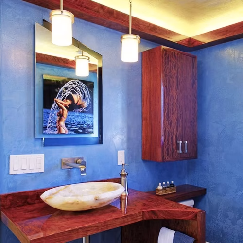 A modern bathroom featuring a blue textured wall, a wooden vanity with a circular sink, and a large mirror reflecting a colorful artwork. The space is illuminated by stylish pendant lights.